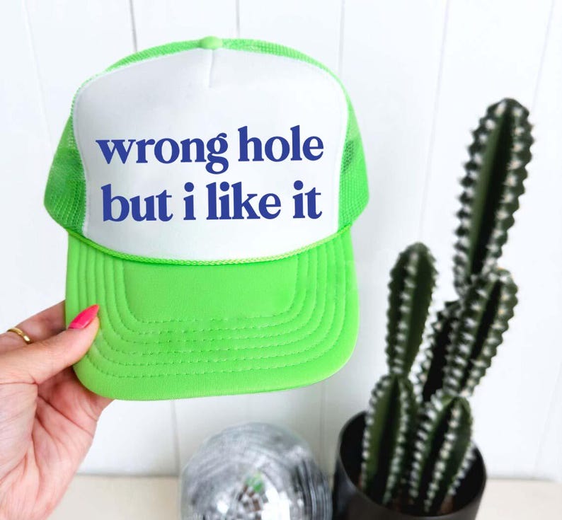 May include: A neon green trucker hat with a white front panel. The text "wrong hole but i like it" is printed in blue on the front. The hat is held by a person's hand. A cactus and disco ball are in the background.