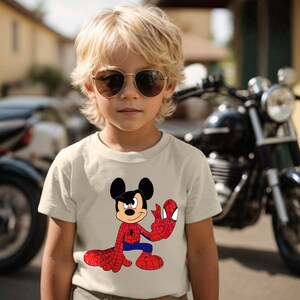 Op de afbeelding: Een beige t-shirt met een stripfiguur die een hybride is van Mickey Mouse en Spider-Man. Het personage draagt een Spider-Man-kostuum. Het kind op de foto draagt een zonnebril en staat voor een motorfiets.