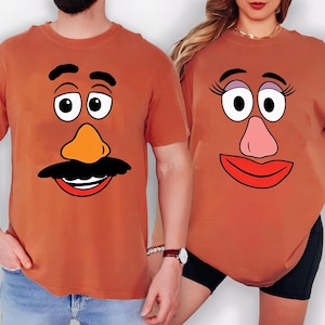 May include: Two rust-colored short-sleeve t-shirts. The left shirt features a cartoon face with black eyebrows, large eyes, an orange nose, and a black mustache over a red mouth. The right shirt features a cartoon face with eyelashes, eyes, a pink nose, and red lips.