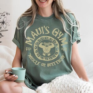 May include: Sage green t-shirt with the text "MAUI'S GYM" in a circular design, featuring a cartoon figure flexing. The phrase "MUSCLE UP, BUTTERCUP" is below the design. The shirt has rolled-up sleeves and a relaxed fit.