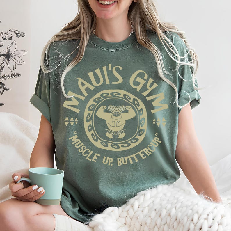 May include: Sage green t-shirt with the text "MAUI'S GYM" in a circular design, featuring a cartoon figure flexing. The phrase "MUSCLE UP, BUTTERCUP" is below the design. The shirt has rolled-up sleeves and a relaxed fit.