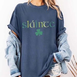 May include: A navy blue t-shirt with the word "Sl&aacute;inte" in green and yellow lettering and a green shamrock. The shirt is part of a St. Patrick's Day collection.
