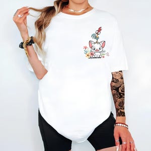 May include: White t-shirt featuring a cartoon pig character with a rooster on its head, surrounded by colorful flowers. The design is on the left chest area. The shirt is paired with black shorts.