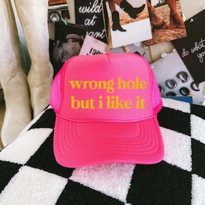 May include: A bright pink trucker hat with the text "wrong hole but i like it" in yellow. The hat has a mesh back and a curved brim. The hat is sitting on a black and white checkered surface.