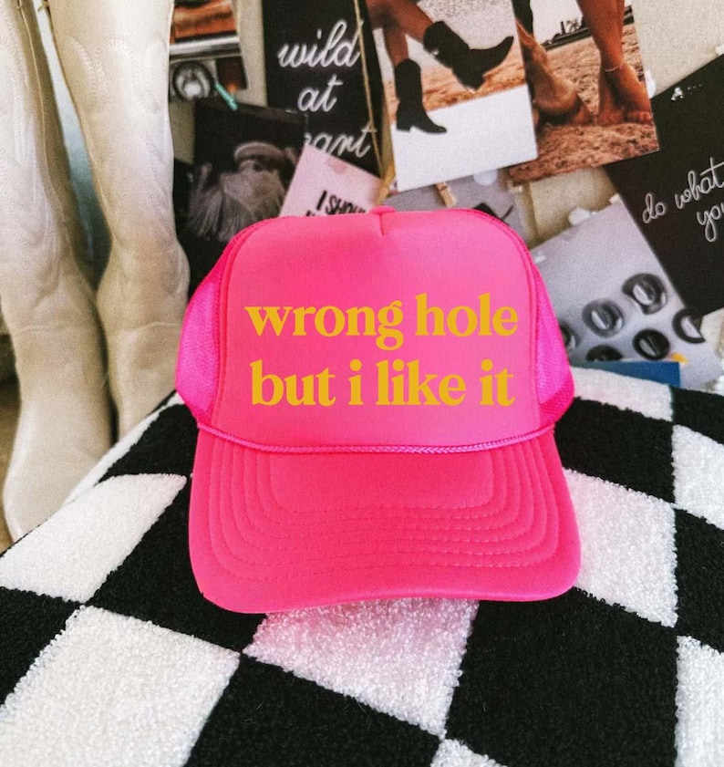 May include: A bright pink trucker hat with the text "wrong hole but i like it" in yellow. The hat has a mesh back and a curved brim. The hat is sitting on a black and white checkered surface.