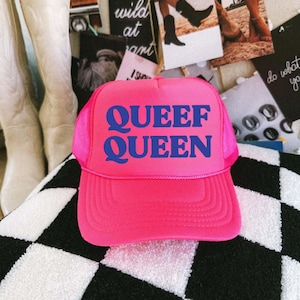 May include: A bright pink trucker hat with the words "QUEEF QUEEN" in bold blue letters. The hat has a mesh back and a curved brim. The hat is sitting on a black and white checkered surface.