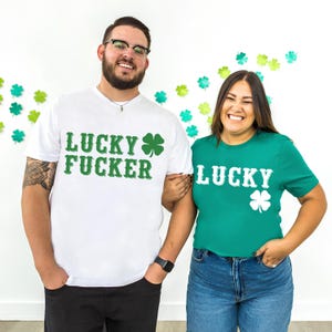 May include: Two people wearing matching St. Patrick's Day t-shirts. One shirt is white with the text "LUCKY FUCKER" and a green shamrock. The other shirt is green with the word "LUCKY" and a white shamrock.