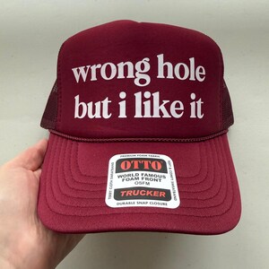 May include: Maroon trucker hat with the text "wrong hole but i like it" in white. The hat has a mesh back and a curved brim. The front of the hat has a foam front and a durable snap closure.
