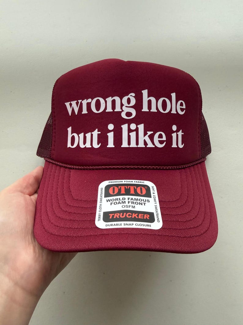 May include: Maroon trucker hat with the text "wrong hole but i like it" in white. The hat has a mesh back and a curved brim. The front of the hat has a foam front and a durable snap closure.