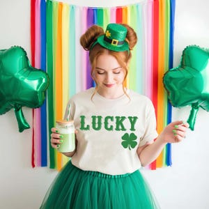May include: A person wearing a cream-colored t-shirt with the word "LUCKY" and a green shamrock graphic. They are also wearing a green tutu and a green glitter top hat. Two green shamrock balloons are on either side.