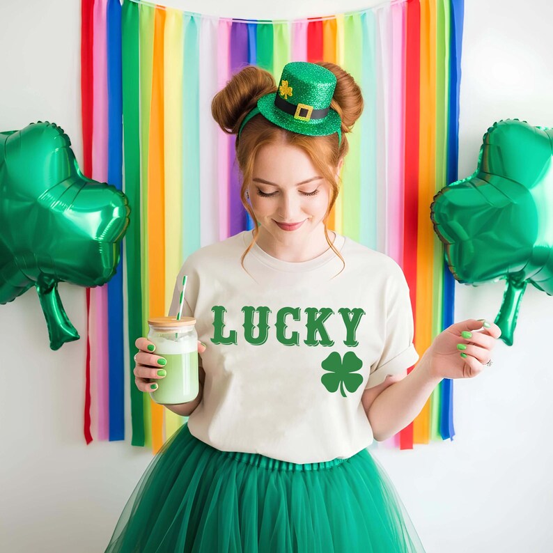 May include: A person wearing a cream-colored t-shirt with the word "LUCKY" and a green shamrock graphic. They are also wearing a green tutu and a green glitter top hat. Two green shamrock balloons are on either side.