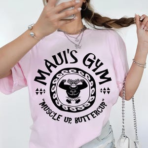 May include: A light pink t-shirt with a black graphic that reads "MAUI'S GYM" in a circular design. The design includes a cartoon figure flexing and the text "MUSCLE UP, BUTTERCUP".