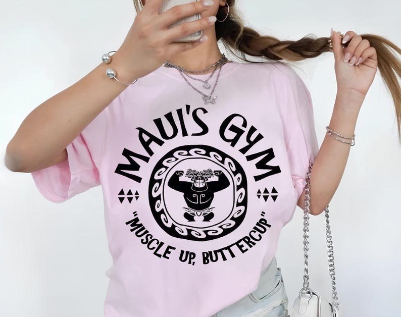 May include: A light pink t-shirt with a black graphic that reads "MAUI'S GYM" in a circular design. The design includes a cartoon figure flexing and the text "MUSCLE UP, BUTTERCUP".