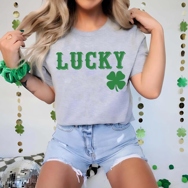 May include: A light gray t-shirt with the word "LUCKY" in green, along with a green four-leaf clover graphic. The shirt is paired with denim shorts. The image has a St. Patrick's Day theme.