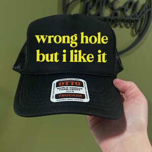 May include: Black trucker hat with the text "wrong hole but i like it" in yellow block letters. The hat has a mesh back and a snap closure. A white tag with the brand name "OTTO" is visible on the front.