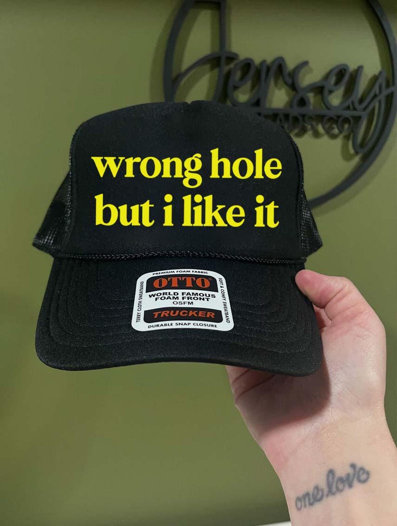 May include: Black trucker hat with the text "wrong hole but i like it" in yellow block letters. The hat has a mesh back and a snap closure. A white tag with the brand name "OTTO" is visible on the front.