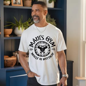 May include: White t-shirt with the text "Maui's Gym" in a circular design, featuring a cartoon weightlifter. The shirt also includes the phrase "Muscle Up Buttercup" in black. The shirt is made of a soft material.