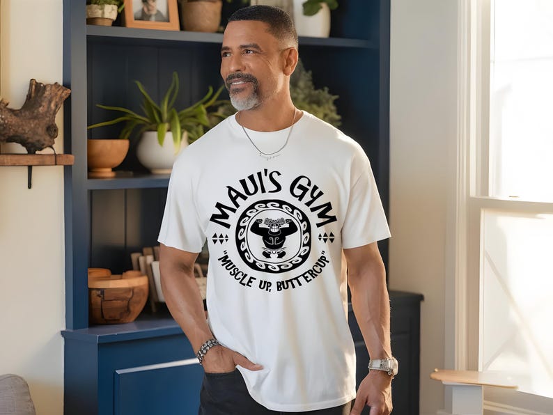 May include: White t-shirt with the text "Maui's Gym" in a circular design, featuring a cartoon weightlifter. The shirt also includes the phrase "Muscle Up Buttercup" in black. The shirt is made of a soft material.
