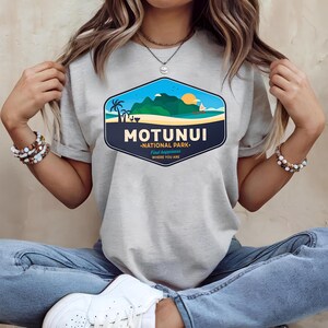 May include: A light grey t-shirt with a colourful graphic depicting a tropical scene. The design includes the text "MOTUNUI - NATIONAL PARK - Find Happiness Where You Are", palm trees, mountains, and a setting sun.