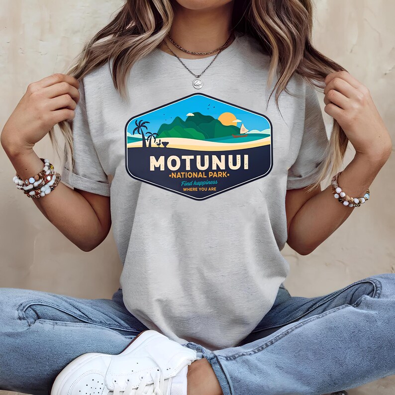 May include: A light grey t-shirt with a colourful graphic depicting a tropical scene. The design includes the text "MOTUNUI - NATIONAL PARK - Find Happiness Where You Are", palm trees, mountains, and a setting sun.