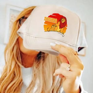 May include: A white trucker hat featuring a graphic of Simba and Mufasa from The Lion King, with the words "Hakuna Matata" in orange. The hat has a mesh back and a curved brim.