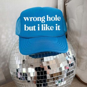 May include: A blue trucker hat with the text "wrong hole but I like it" in white. The hat is resting on a mirrored disco ball. The hat has a mesh back and a curved brim.