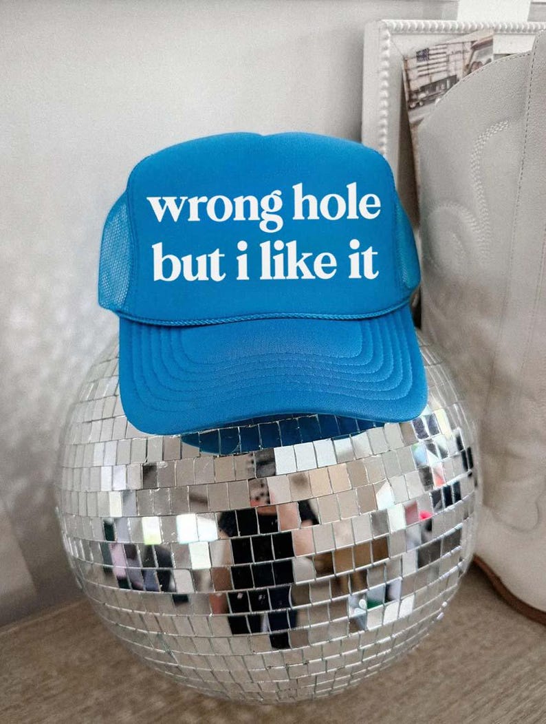 May include: A blue trucker hat with the text "wrong hole but I like it" in white. The hat is resting on a mirrored disco ball. The hat has a mesh back and a curved brim.
