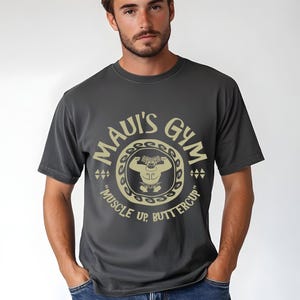 May include: Dark gray t-shirt with a circular graphic that reads "MAUI'S GYM" in a gold color. The graphic includes a weightlifter and the text "MUSCLE UP, BUTTERCUP". The shirt is a crew neck.
