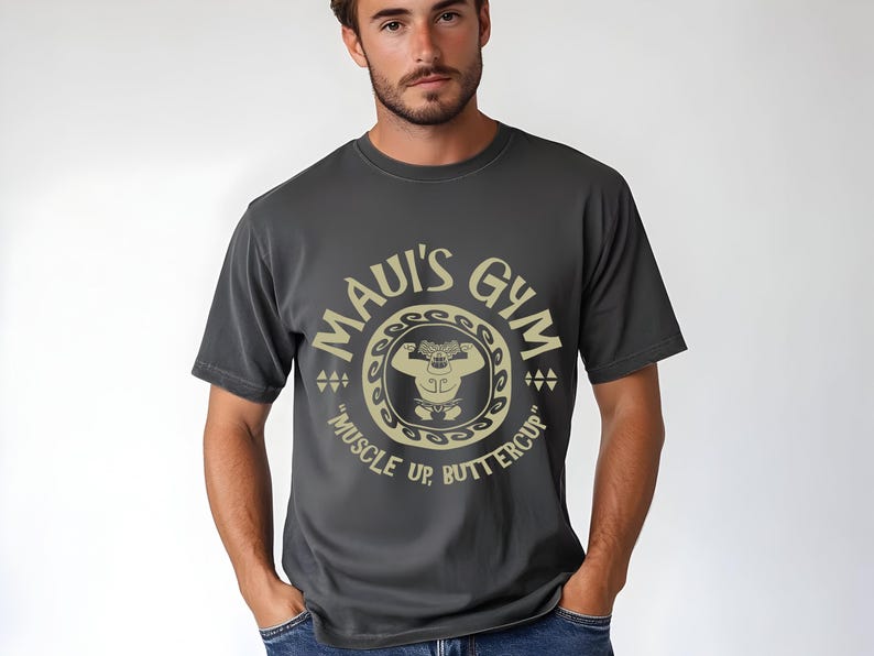 May include: Dark gray t-shirt with a circular graphic that reads "MAUI'S GYM" in a gold color. The graphic includes a weightlifter and the text "MUSCLE UP, BUTTERCUP". The shirt is a crew neck.