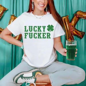 May include: White t-shirt with the text "LUCKY FUCKER" in green, along with a green shamrock. The person is holding a glass of green beer. Gold foil letter balloons spell out the letter "K".