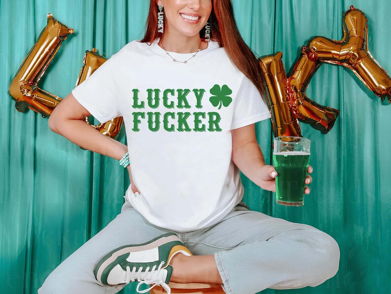 May include: White t-shirt with the text "LUCKY FUCKER" in green, along with a green shamrock. The person is holding a glass of green beer. Gold foil letter balloons spell out the letter "K".