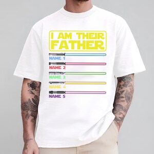 May include: White t-shirt with the text "I AM THEIR FATHER" in yellow. Below the text are five lightsabers with different colored blades and the text "NAME" followed by a number. The shirt is a crew neck.