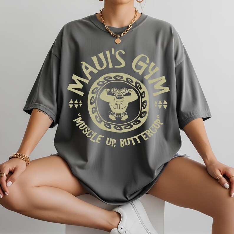 May include: Gray oversized t-shirt with a gold graphic that reads "Maui's Gym" with a cartoon figure flexing. The text "Muscle Up, Buttercup" is below the graphic. The shirt has short sleeves and a crew neck.