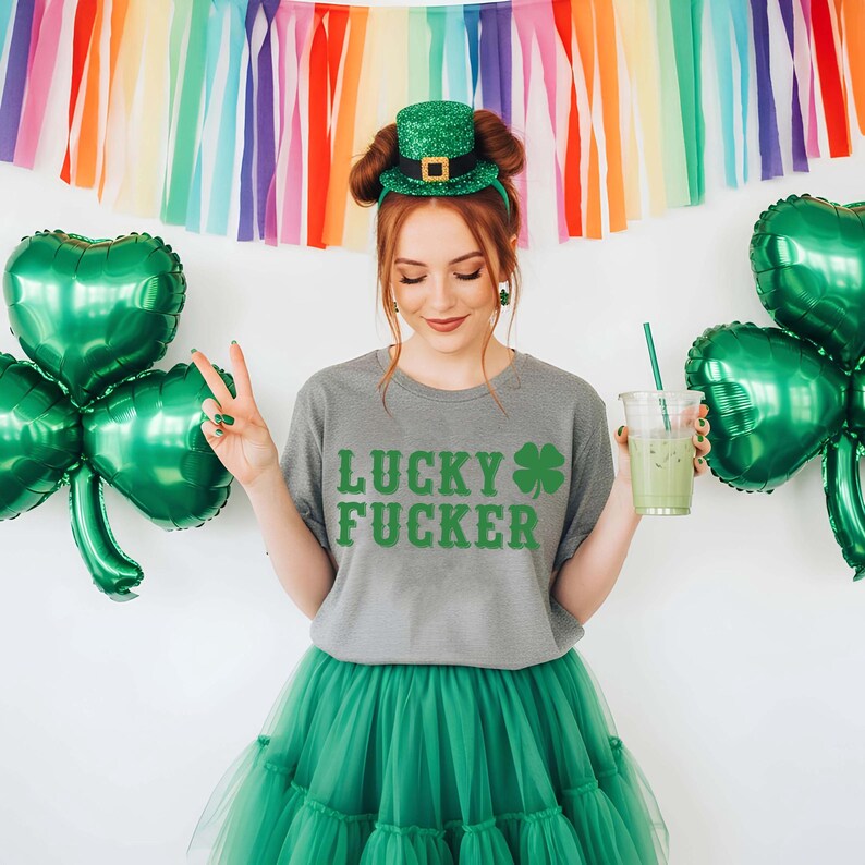 May include: A person wearing a gray t-shirt with the words "LUCKY FUCKER" in green, paired with a green tutu and a green glitter top hat. Green shamrock balloons and a rainbow streamer backdrop complete the St. Patrick's Day theme.