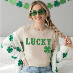 May include: A beige t-shirt with the word "LUCKY" in green, and a green shamrock graphic. The person is wearing green shamrock-shaped sunglasses and a white cardigan with green shamrock designs. A green shamrock garland hangs in the background.