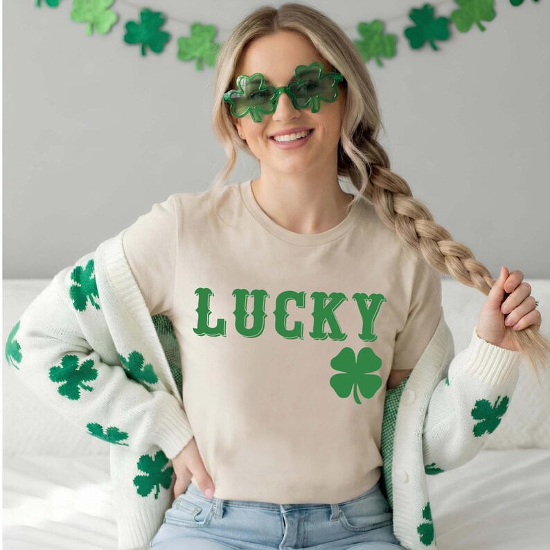 May include: A beige t-shirt with the word "LUCKY" in green, and a green shamrock graphic. The person is wearing green shamrock-shaped sunglasses and a white cardigan with green shamrock designs. A green shamrock garland hangs in the background.
