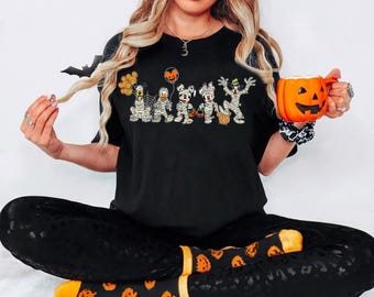 Könnte beinhalten: Ein schwarzes T-Shirt mit einem Motiv von Mickey Mouse und seinen Freunden, die als Mumien für Halloween verkleidet sind. Die Figuren halten Kürbisse und Süßigkeiten. Das T-Shirt wird von einer Person mit langen blonden Haaren getragen, die eine Kürbis-Tasse in der Hand hält.