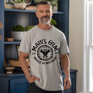 May include: A light gray t-shirt with the text "MAUI'S GYM" in a circular design, featuring a weightlifter graphic. The shirt also includes the phrase "MUSCLE UP, BUTTERCUP" in black. The person is wearing a silver chain and bracelet.
