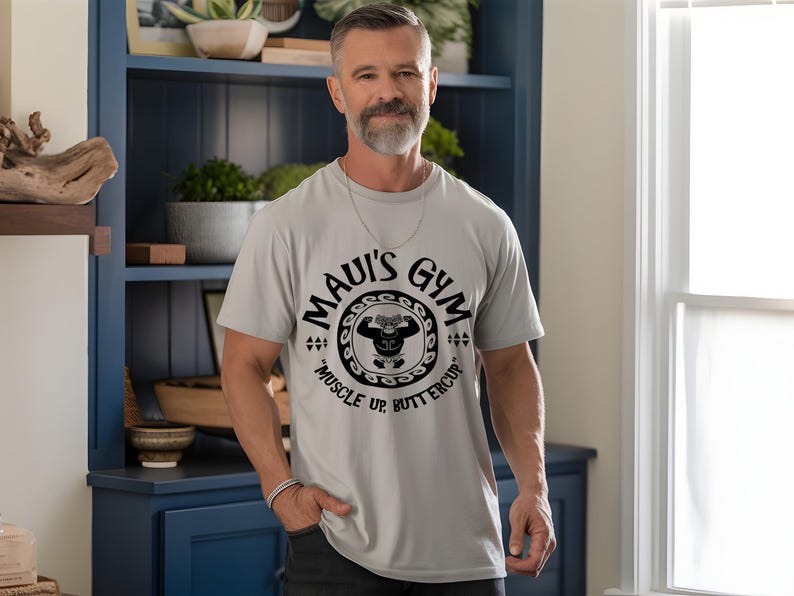 May include: A light gray t-shirt with the text "MAUI'S GYM" in a circular design, featuring a weightlifter graphic. The shirt also includes the phrase "MUSCLE UP, BUTTERCUP" in black. The person is wearing a silver chain and bracelet.