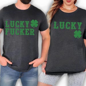 May include: Two dark gray t-shirts with green text and a green shamrock. One shirt says "LUCKY FUCKER" and the other says "LUCKY". The shirts are short-sleeved and appear to be made of a soft material.