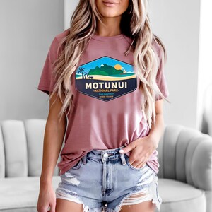 May include: Mauve t-shirt with a tropical landscape graphic, including palm trees, mountains, and a setting sun, and the text "MOTUNUI NATIONAL PARK". The shirt is worn with denim shorts. The design is in a hexagonal shape.