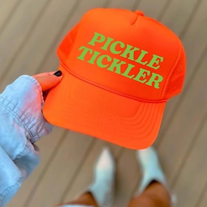 May include: Bright orange trucker hat with the words "PICKLE TICKLER" in green block letters. The hat has a mesh back and a curved brim. A fun, casual accessory.