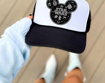 May include: A black and white trucker hat featuring a graphic of a Mickey Mouse silhouette filled with Star Wars icons and the text Star Wars. The hat has a white front panel, a black brim, and a black mesh back panel with an adjustable snap closure.