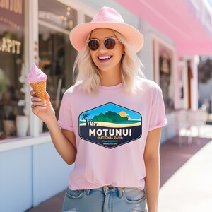 May include: A pale pink t-shirt featuring a tropical island graphic with palm trees, mountains, and a sailboat, and the text "MOTUNUI - NATIONAL PARK". The shirt is worn with a pink hat and sunglasses.