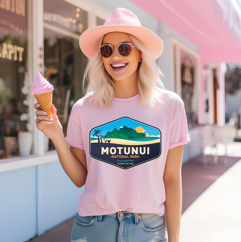 May include: A pale pink t-shirt featuring a tropical island graphic with palm trees, mountains, and a sailboat, and the text "MOTUNUI - NATIONAL PARK". The shirt is worn with a pink hat and sunglasses.