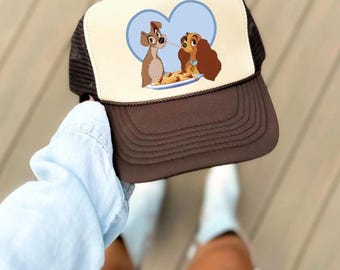 May include: A brown and tan trucker hat featuring a cartoon image of two dogs sharing spaghetti, set against a light blue heart-shaped background. The hat has a brown brim and a mesh back.