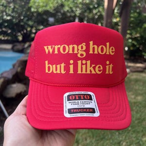 May include: Red trucker hat with the text "wrong hole but i like it" in yellow. The hat has a foam front and a mesh back. The hat also has a "TRUCKER" label.