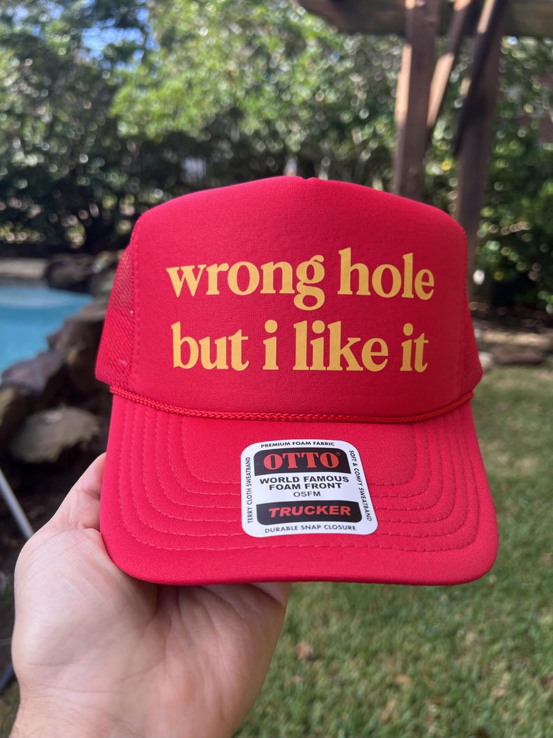 May include: Red trucker hat with the text "wrong hole but i like it" in yellow. The hat has a foam front and a mesh back. The hat also has a "TRUCKER" label.