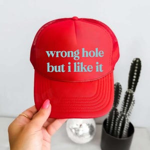 May include: A red trucker hat with the text "wrong hole but i like it" in turquoise. The hat has a mesh back and a curved brim. The hat is being held by a person.