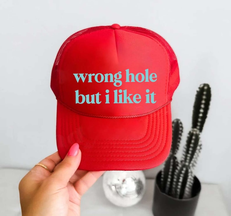 May include: A red trucker hat with the text "wrong hole but i like it" in turquoise. The hat has a mesh back and a curved brim. The hat is being held by a person.